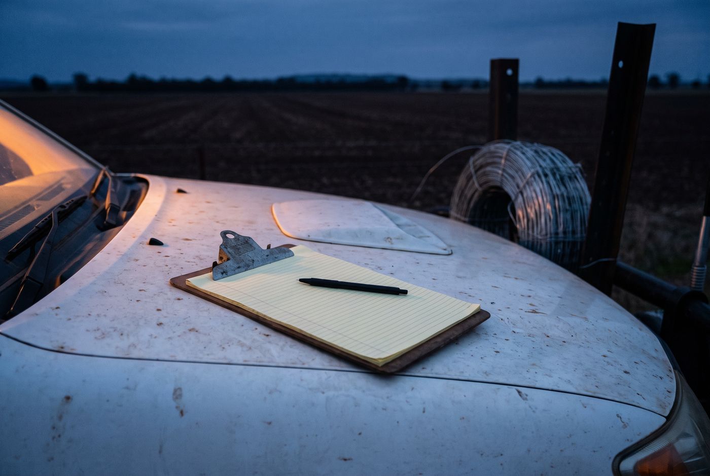 A clipboard and pen resting on the bonnet of a ute at dusk, a roll of wire and a strainer post behind